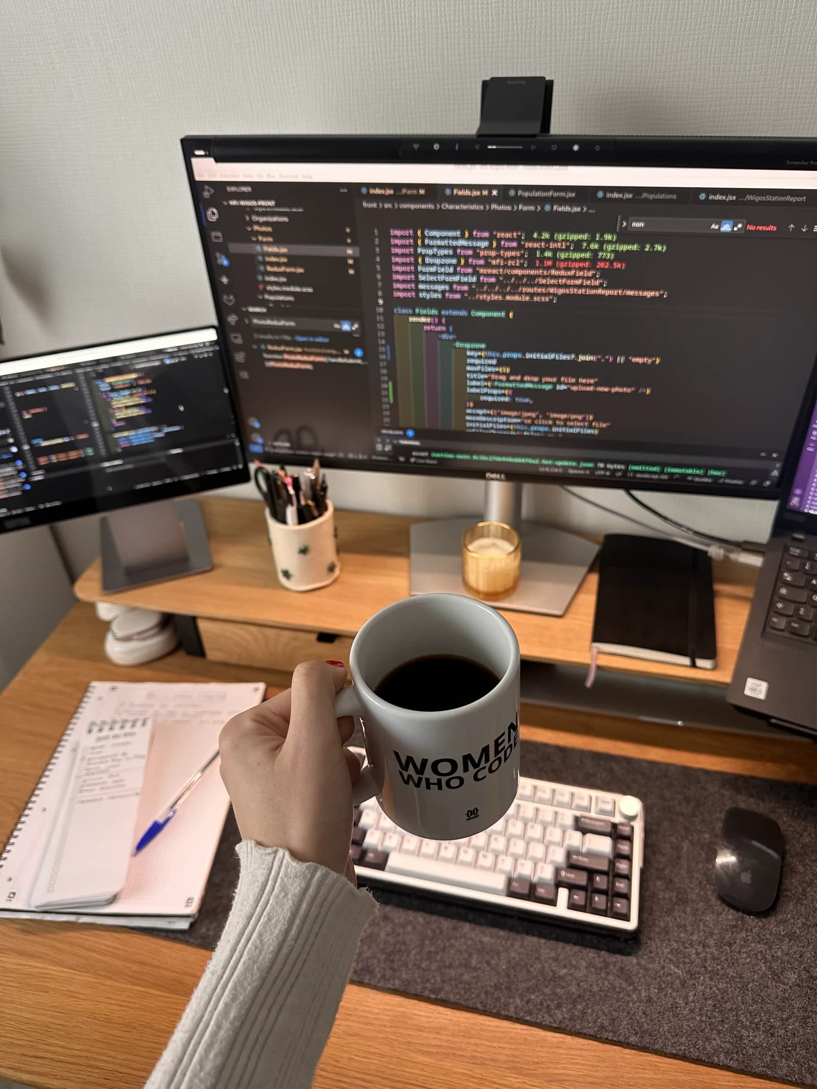 A hand holding a 'Women Who Code' mug in front of a monitor showing React code